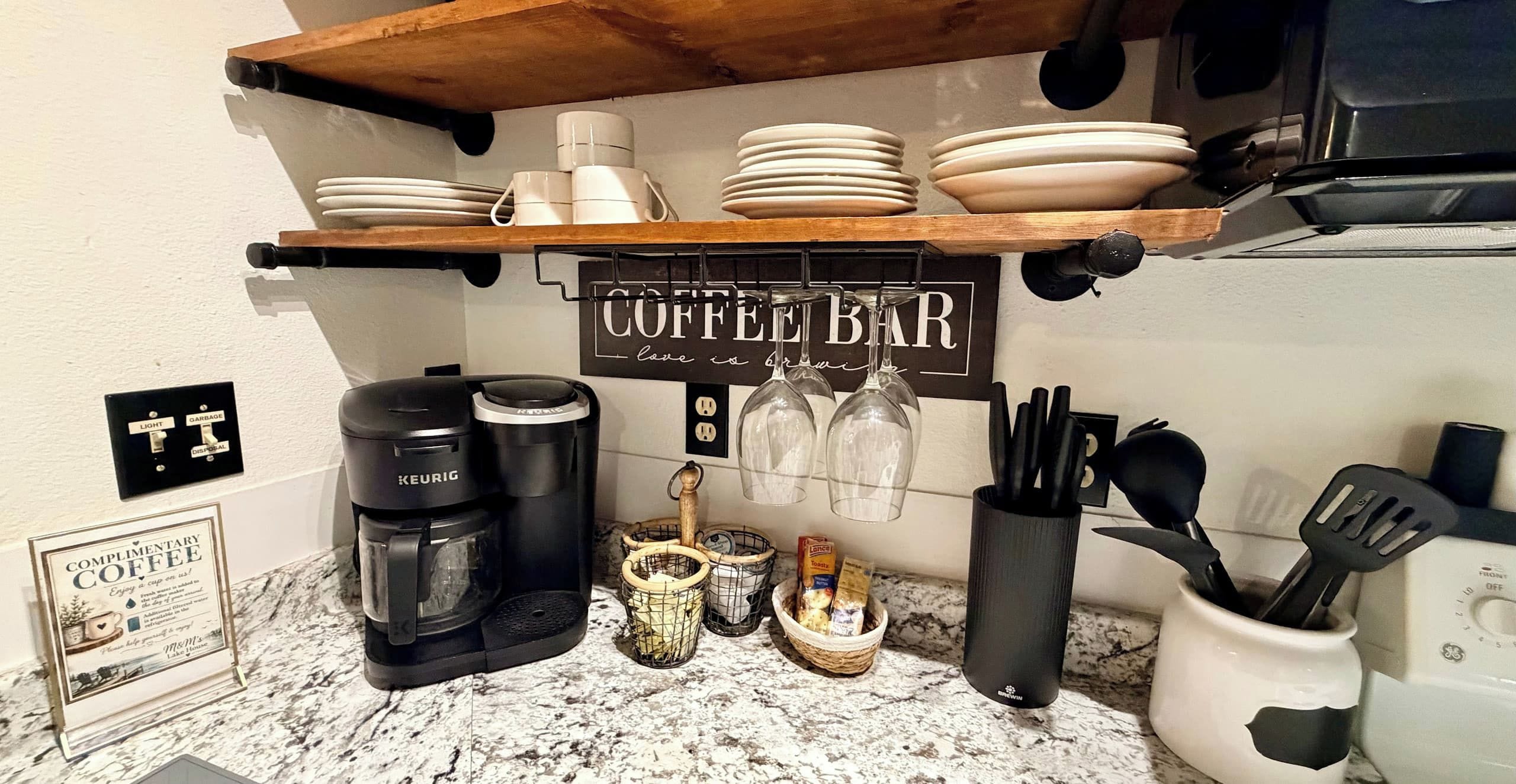 Uma bancada de cozinha com cafeteira, moedor de café, cesta de grãos de café, xícara de café e placa indicando 'coffee bar'.