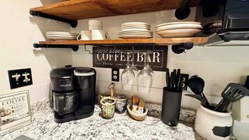 Uma bancada de cozinha com cafeteira, moedor de café, cesta de grãos de café, xícara de café e placa indicando 'coffee bar'.