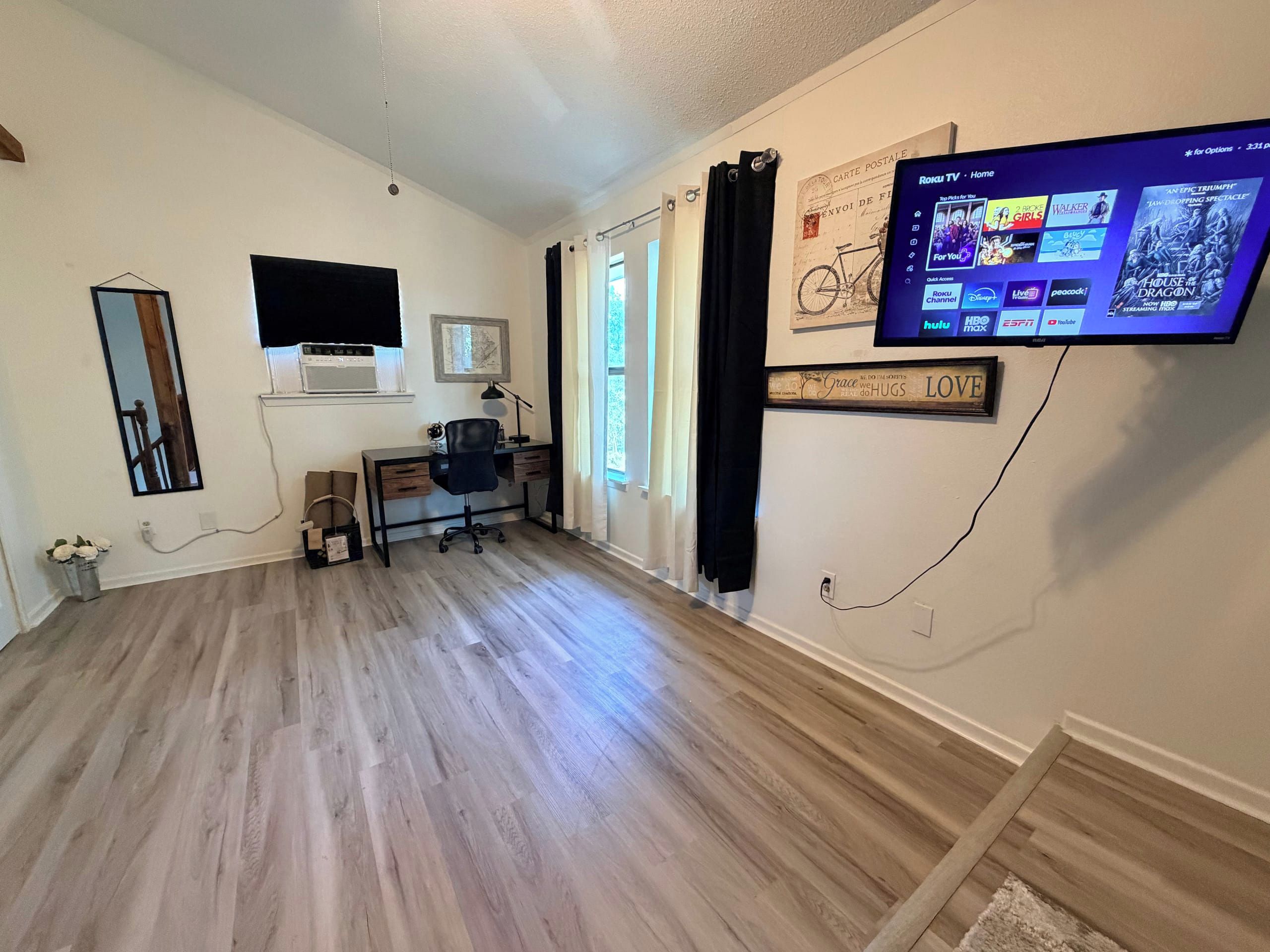 Un salon doté d’un téléviseur à écran plat fixé au mur, d’un bureau avec une chaise et d’une grande fenêtre avec des rideaux.