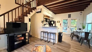 A spacious living room with a wooden staircase, a television, a coffee table, and a kitchen area with a bar.