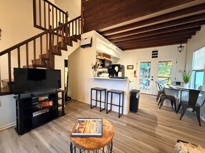 Uma sala de estar ampla, com escada de madeira, televisão, mesa de centro e área de cozinha com balcão.