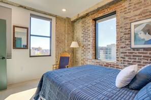A bedroom with a large bed, a chair, a window, and a brick wall. - 7th-floor Condo: Walk to Cafes, Dining & More (Chattanooga)