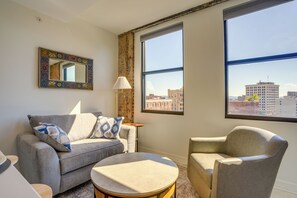 A living room with a sofa, armchair, and a round coffee table. There is a large window with a view of buildings. - 7th-floor Condo: Walk to Cafes, Dining & More (Chattanooga)