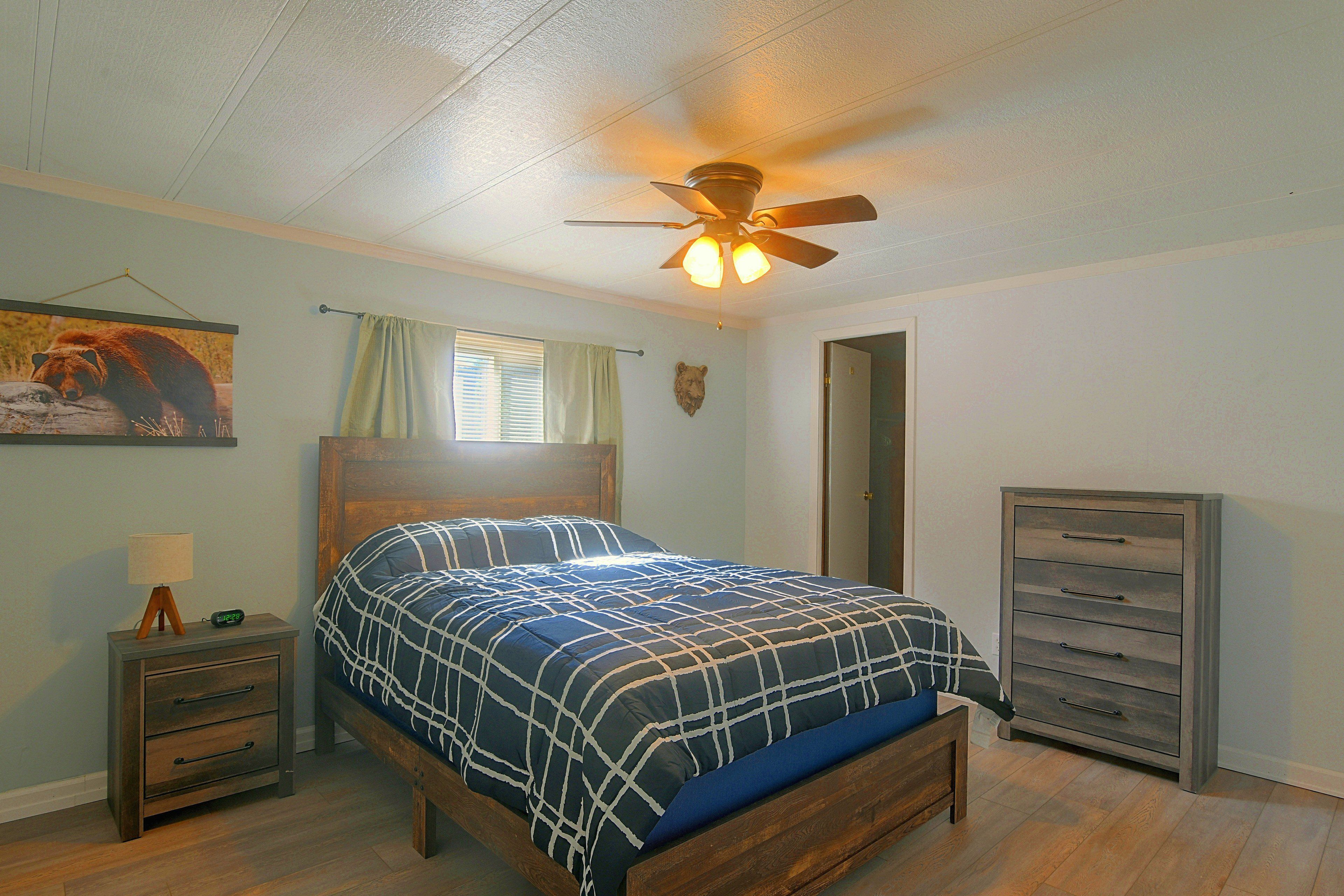 A bedroom with a bed, a nightstand, a dresser, a ceiling fan, a window with blinds, and a bear-themed wall decoration.