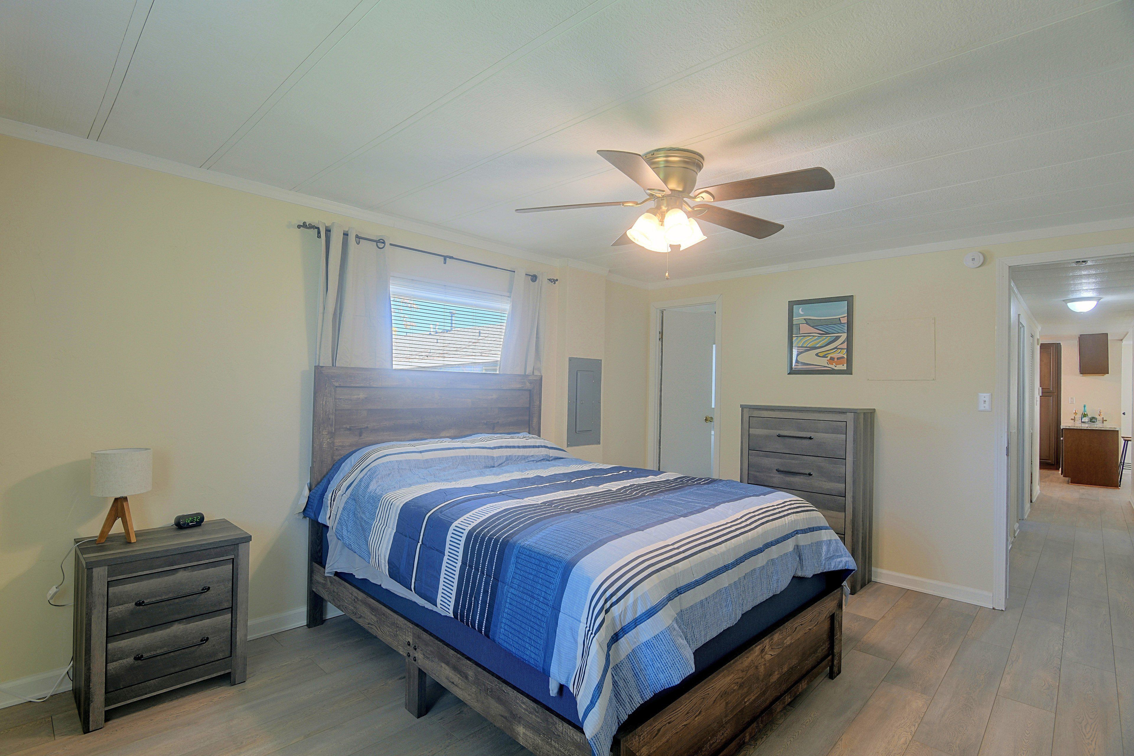Une chambre à coucher comprenant un lit, une table de chevet, un ventilateur de plafond, une fenêtre avec des rideaux et une porte donnant sur une autre pièce.