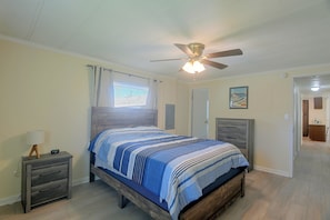 Ein Schlafzimmer mit einem Bett, einem Nachttisch, einem Deckenventilator, einem Fenster mit Vorhängen und einer Tür, die in einen anderen Raum führt.