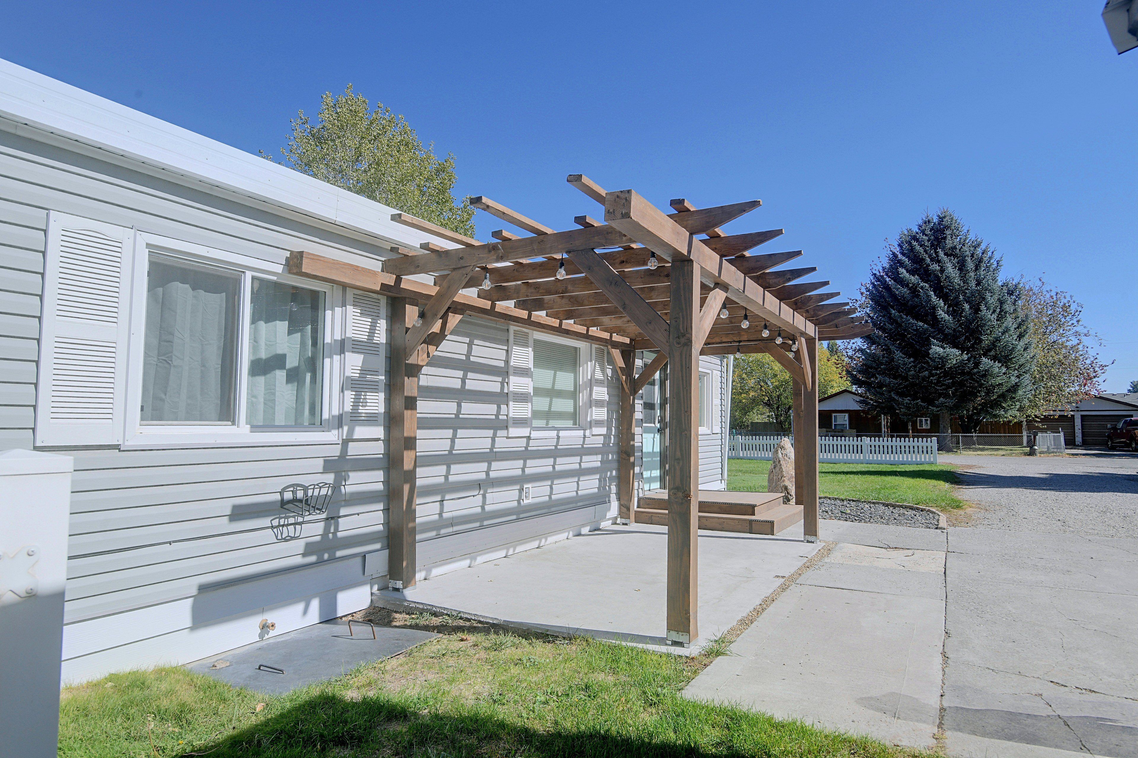 Une structure de pergola en bois, reposant sur des fondations en béton, jouxtant une maison mobile blanche dotée d’une fenêtre et d’une porte.