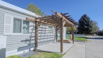 Une structure de pergola en bois, reposant sur des fondations en béton, jouxtant une maison mobile blanche dotée d’une fenêtre et d’une porte.