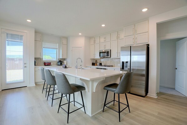A modern kitchen with a central island, bar stools, stainless steel appliances, and white cabinetry. - 7 Mi to Dtwn Rosenberg: Spacious Family Home (Rosenberg)