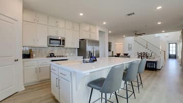 A modern kitchen with white cabinetry, a central island with bar stools, and a dining area with a staircase leading to another level.