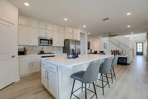 A modern kitchen with white cabinetry, a central island with bar stools, and a dining area with a staircase leading to another level. - 7 Mi to Dtwn Rosenberg: Spacious Family Home (Rosenberg)