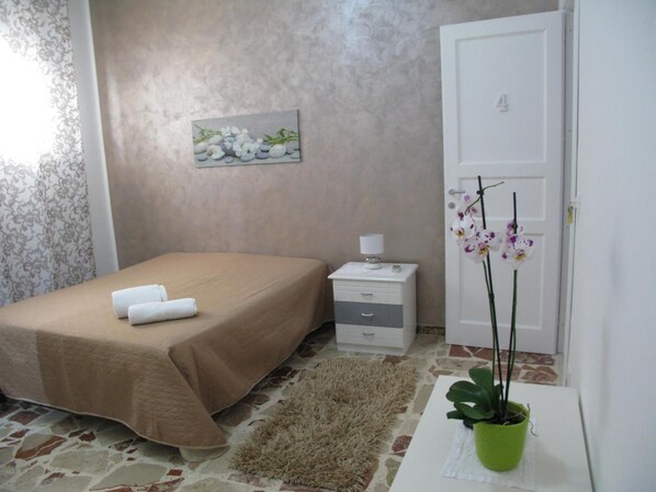 Un dormitorio con cama, mesita de noche, lámpara, un cuadro, una puerta y una planta en maceta.