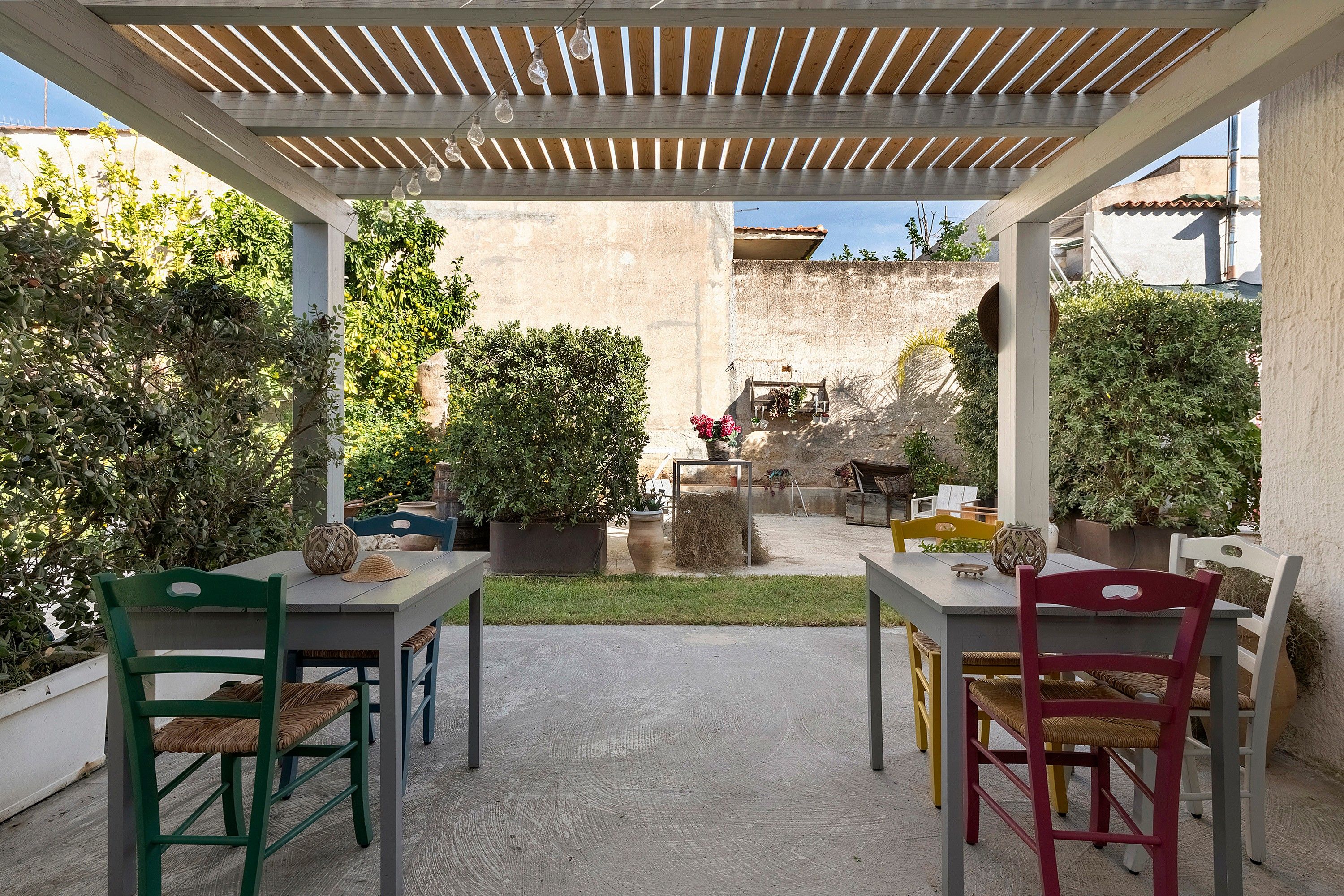 Ein überdachter Patio-Bereich mit zwei Tischen und Stühlen, umgeben von Kübelpflanzen und Blick auf einen Innenhof.
