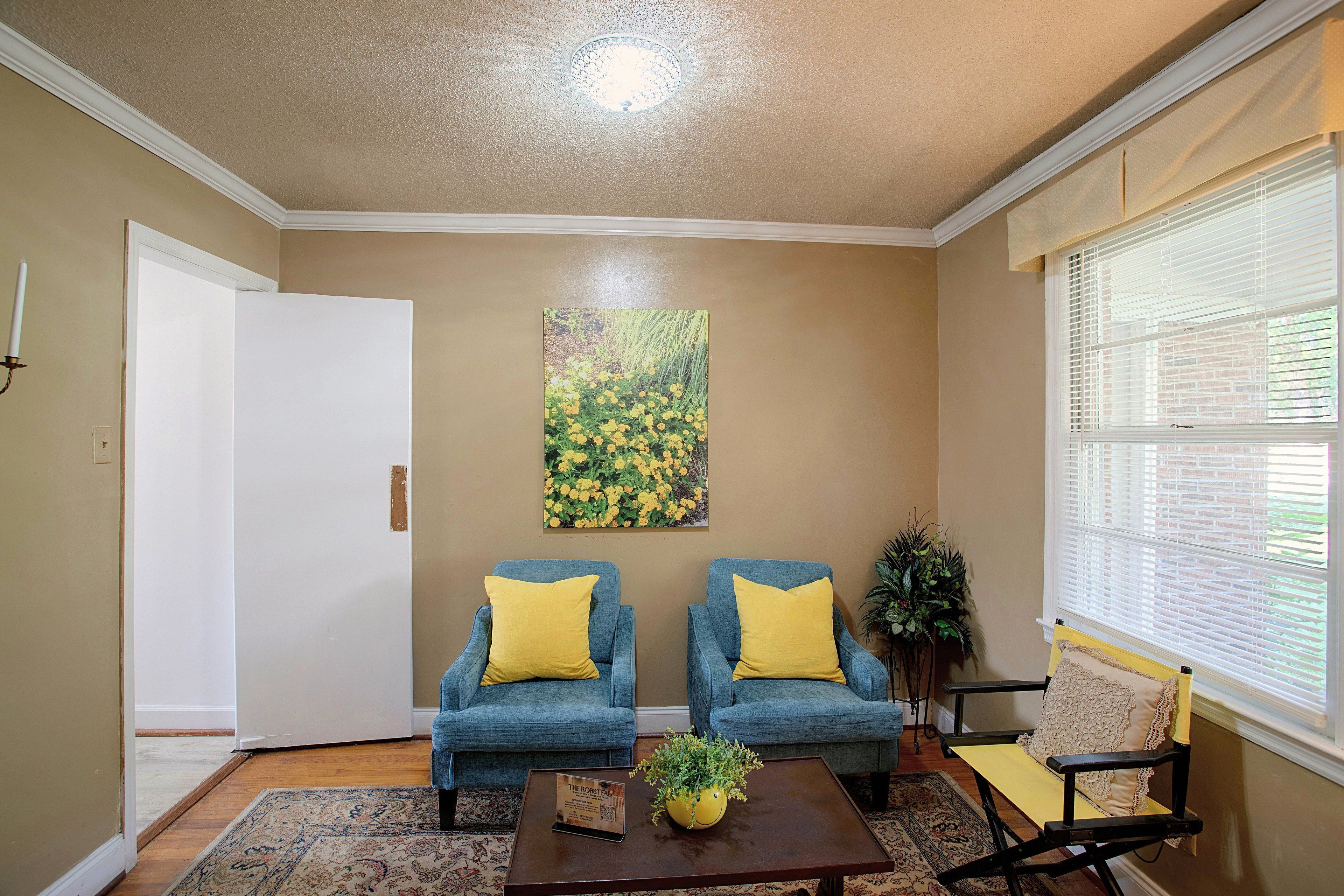A living room with two blue armchairs, a coffee table, a painting, and a window with blinds.