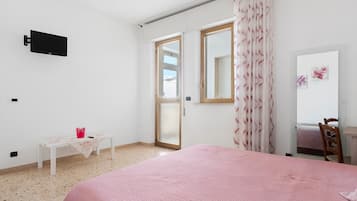 A bedroom with a bed, a TV mounted on the wall, a small table with a vase, and a window with curtains.