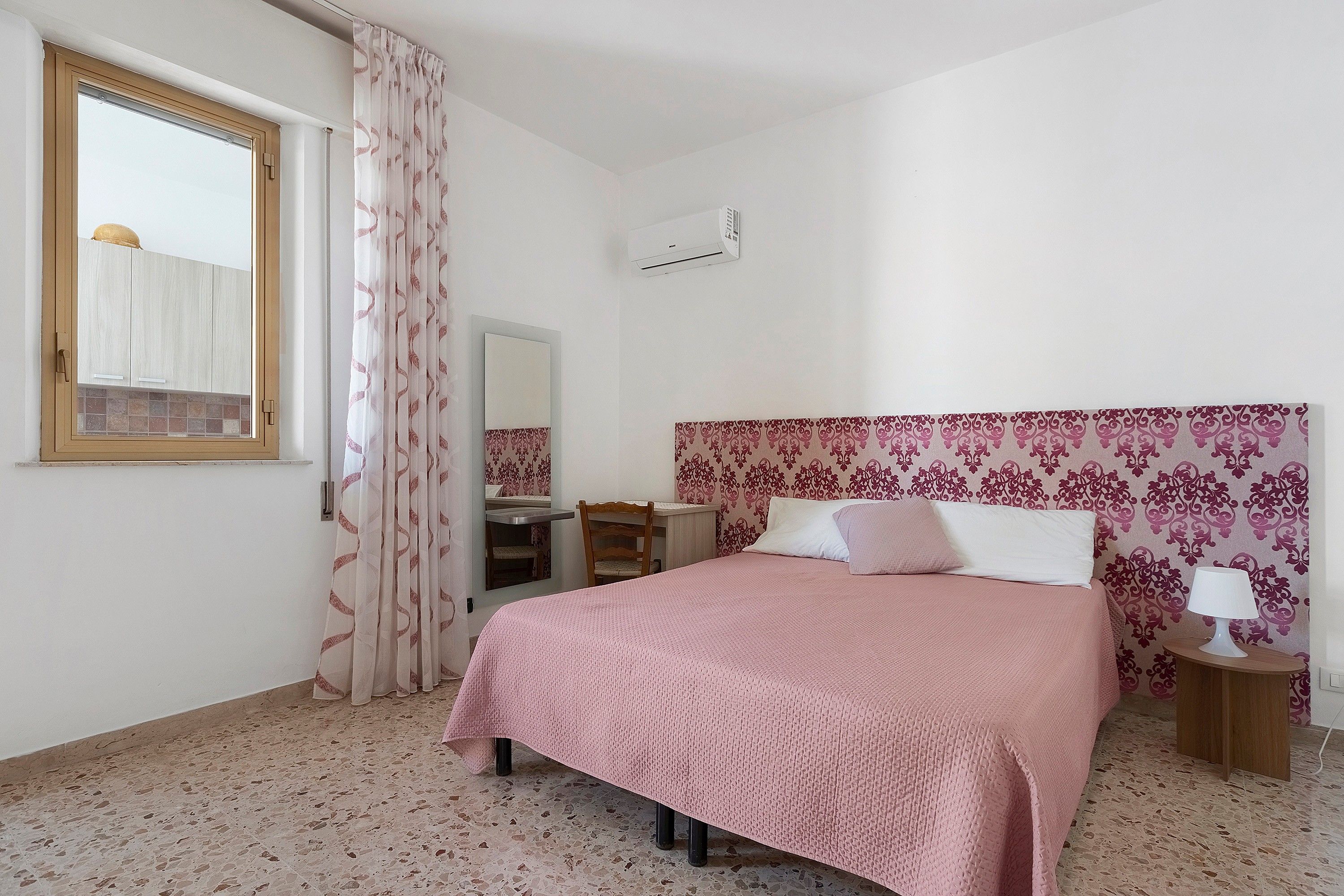 Une chambre à coucher avec un lit, une table de chevet avec une lampe, une fenêtre avec des rideaux, un miroir et un climatiseur fixé au mur.