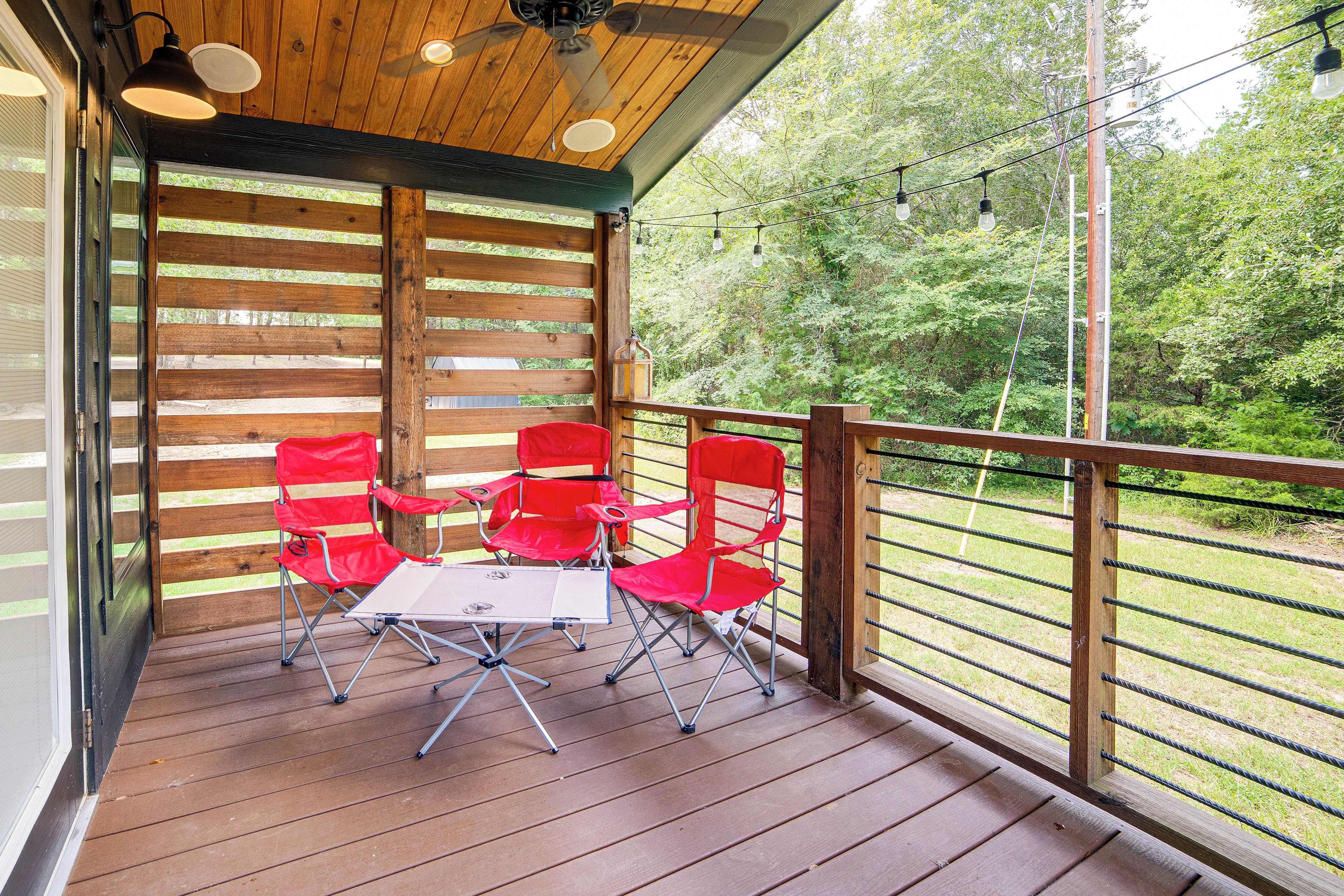 A wooden deck with a table and red chairs, surrounded by a metal railing and a view of greenery.