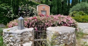 En stengrund mur med en metalport, omgivet af frodige buske med lyserøde og hvide blomster, og en bygning med et dekorativt våbenskjold i baggrunden.