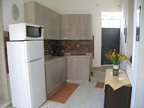 Et lille køkken med mikroovn, komfur og vask. Der er et ur på væggen og en vase med blomster på bordet.