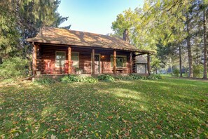 A log cabin with a porch, surrounded by trees and a grassy area with fallen leaves. - Waterfront Log Cabin w/ Fire Pit in Mayville! (Mayville)