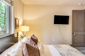 Quarto de hotel com cama, televisão na parede, porta e janela vista para árvores.