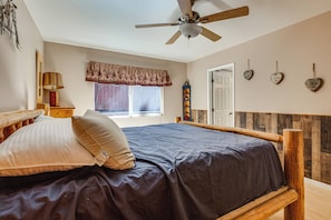 Ein Schlafzimmer mit Bett, Deckenventilator, Fenster mit Vorhängen, Nachttisch mit Lampe und dekorativen herzförmigen Objekten an der Wand.