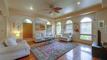 A spacious living room with a large window, a ceiling fan, and a flat-screen TV.