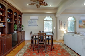 A dining area with a wooden table and chairs, a decorative rug, and a bookshelf with various items.