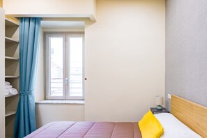 A bedroom with a bed, a window, a nightstand with a lamp, and a shelf with folded towels. - Rega 37 (Naples)