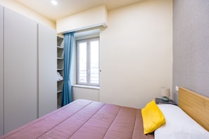 Ein Schlafzimmer mit einem Bett, einem Kleiderschrank, einem Fenster mit Vorhängen, einem Nachttisch mit Lampe und Blick nach außen.