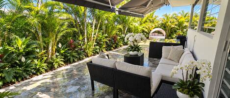 An outdoor patio with a sofa and table, surrounded by lush tropical plants and a clear view of the sky.