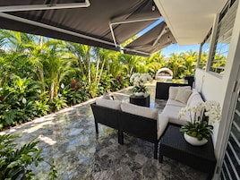 Un patio exterior con sofá y mesa, rodeado de exuberante vegetación tropical y una vista despejada del cielo.