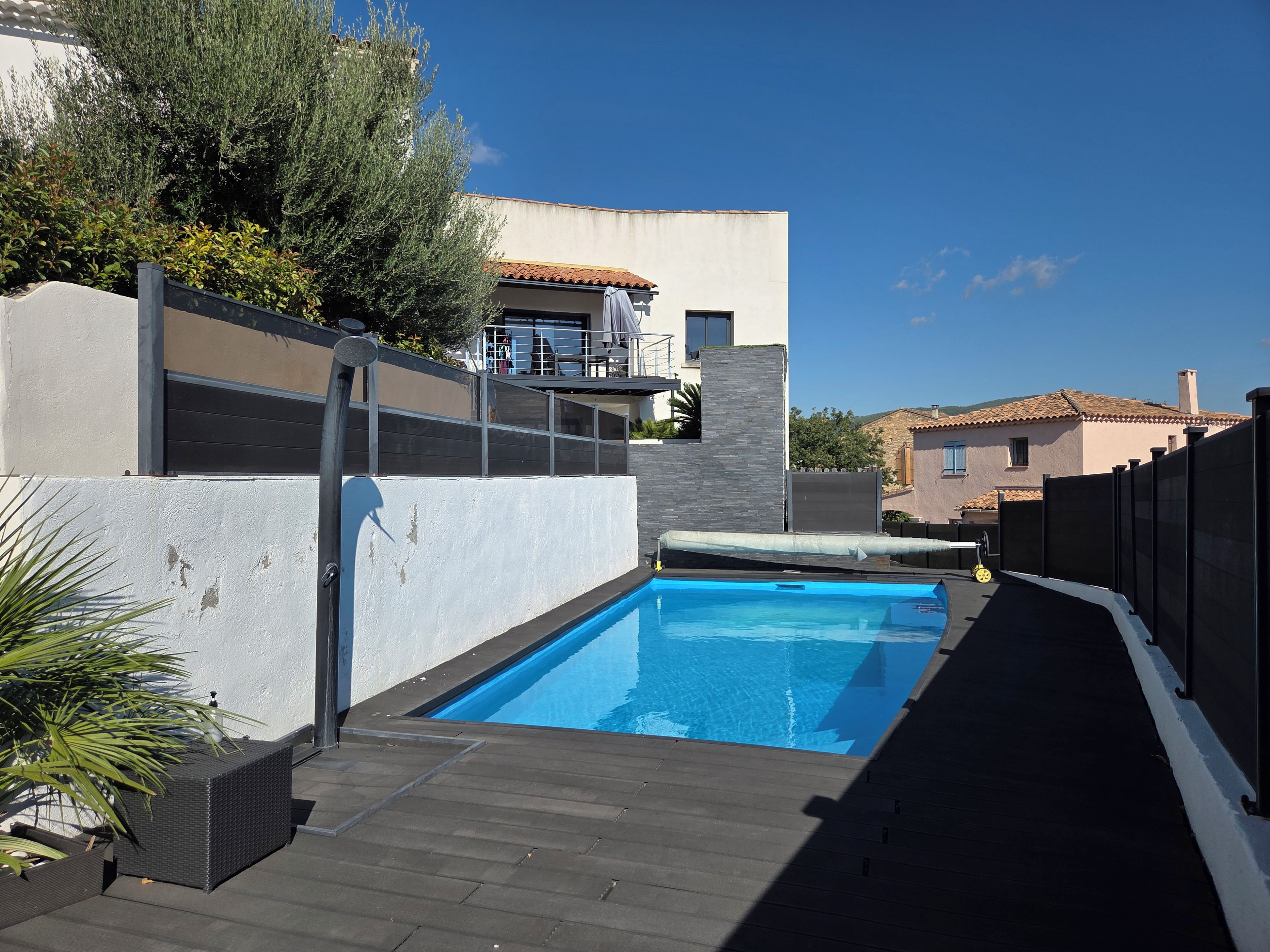 Une piscine avec une terrasse en bois, un mur blanc et, au fond, un bâtiment doté d’un toit recouvert de tuiles.