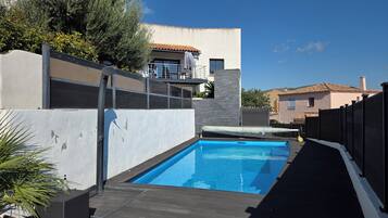 Une piscine avec une terrasse en bois, un mur blanc et, au fond, un bâtiment doté d’un toit recouvert de tuiles.