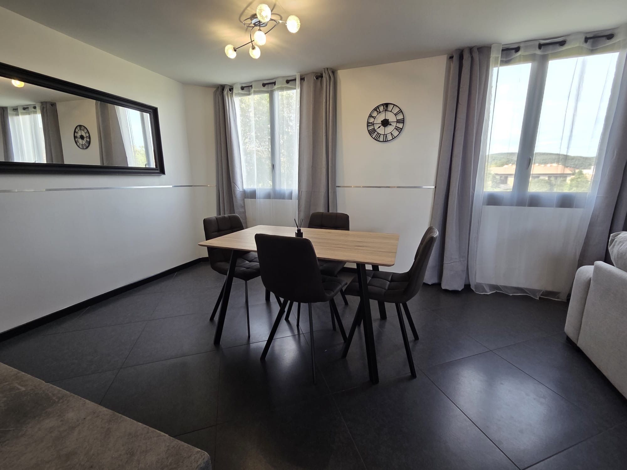 Un espace repas moderne avec une table en bois et des chaises noires, une horloge murale et une grande fenêtre avec des rideaux.