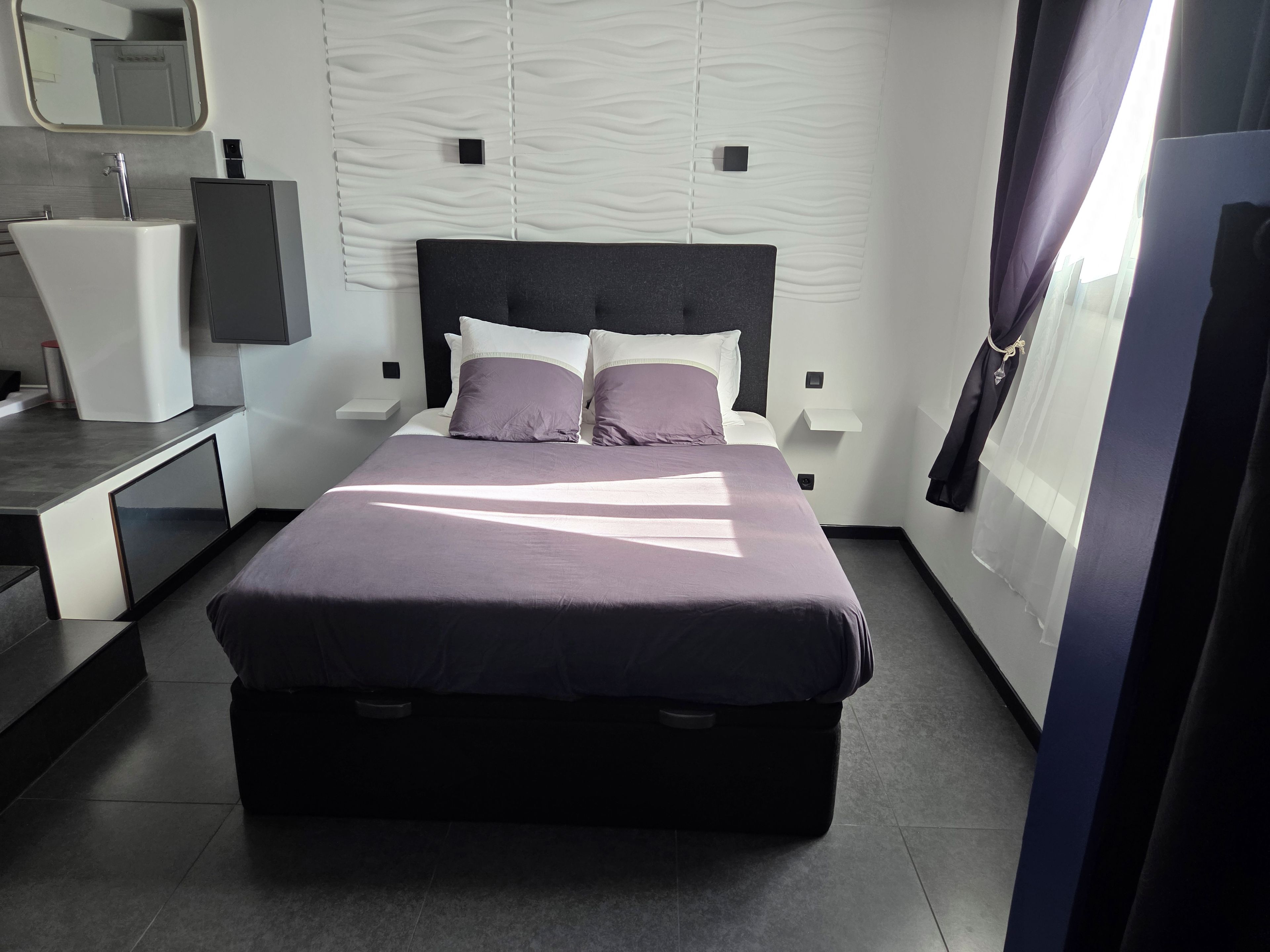 Een moderne slaapkamer met een groot bed, een nachtkastje, een ingebouwde bank, een wandtelevisie en een badkamer met een wastafel en spiegel.