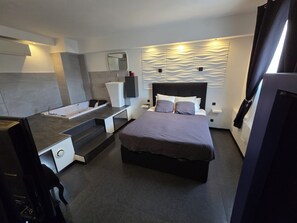 A modern bedroom with a bed, a vanity with a mirror, and a built-in bathtub.