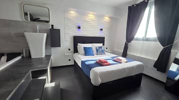 A modern hotel room with a large bed, a bedside table, a mirror, and a wall-mounted light fixture.