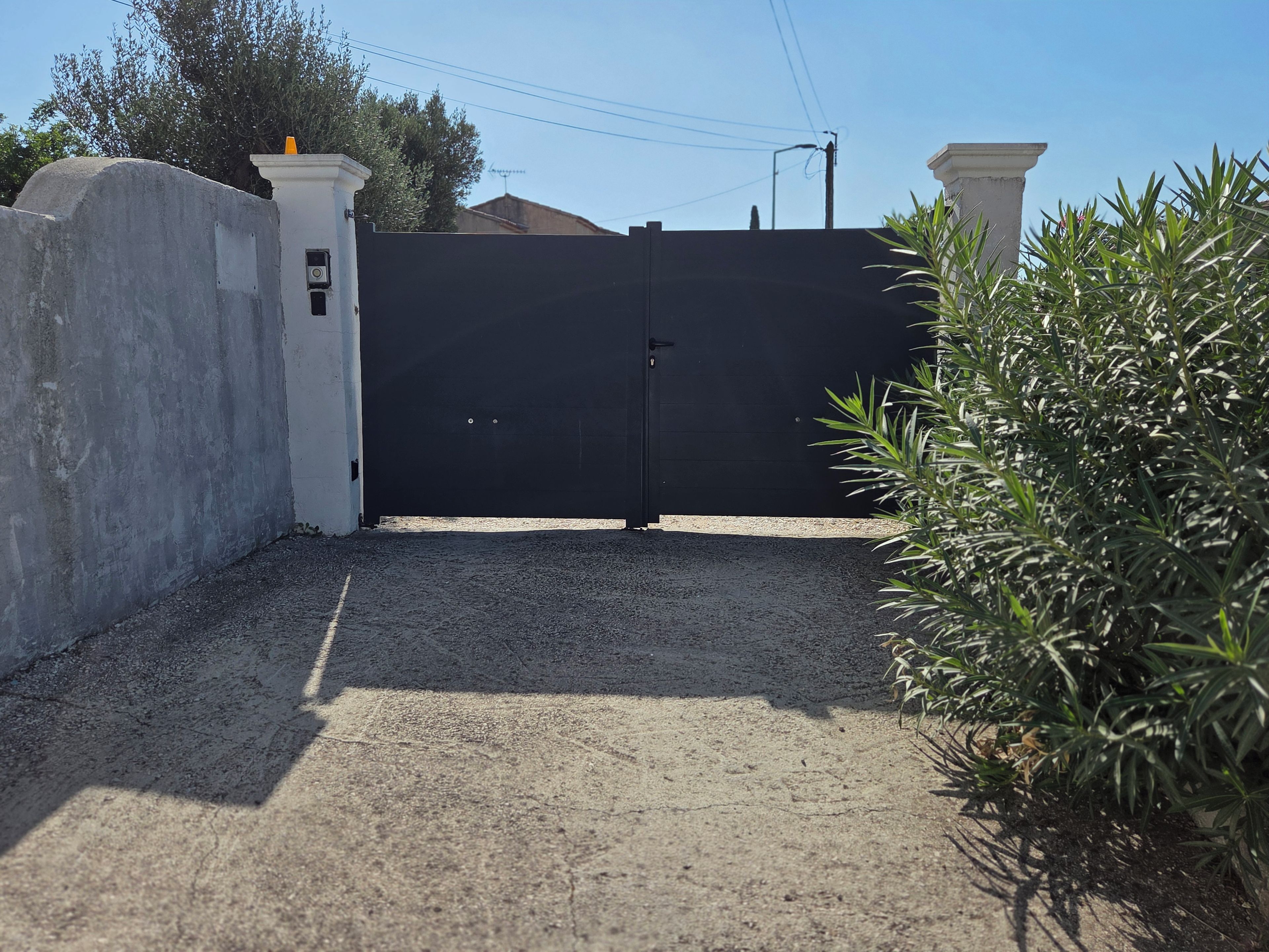 Une entrée fermée par un portail, avec un mur en béton et un portail noir, le tout entouré de gravier et de verdure.