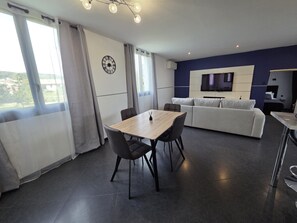 Een moderne woonkamer met een eethoek, een bank, een televisie en uitzicht door grote ramen.