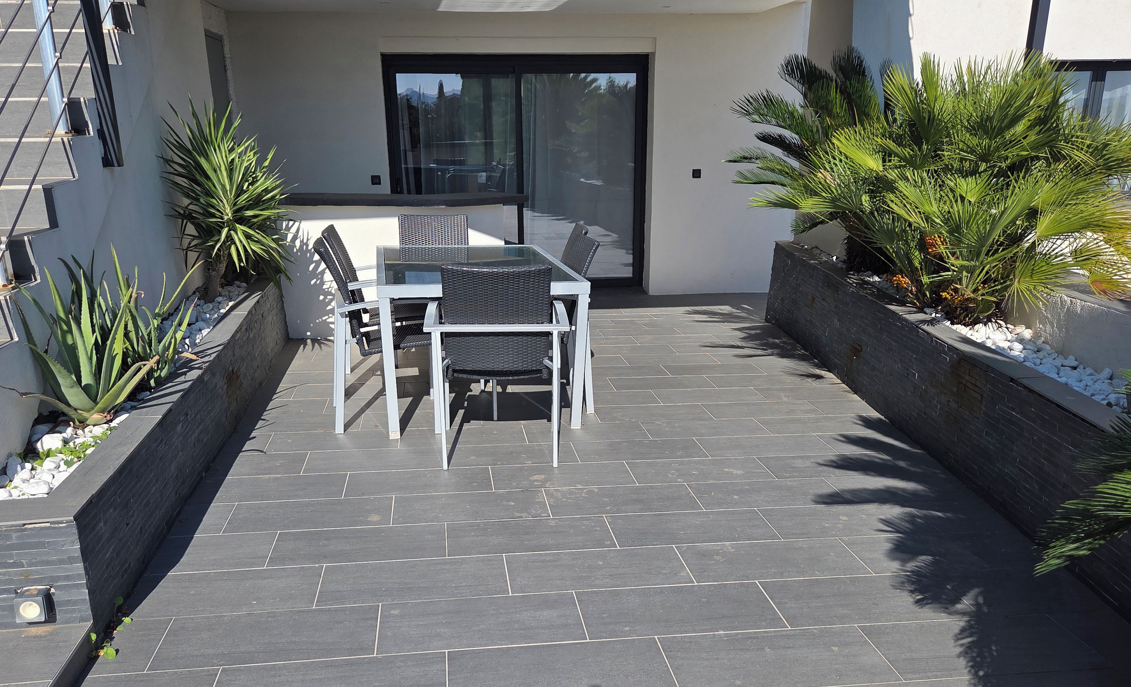 Een terras buiten met een tafel en stoelen, omringd door planten en een stenen vloer.