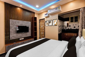 A hotel room with a bed, a flat-screen TV mounted on the wall, a wardrobe, a bathroom with a shower, and a kitchenette.