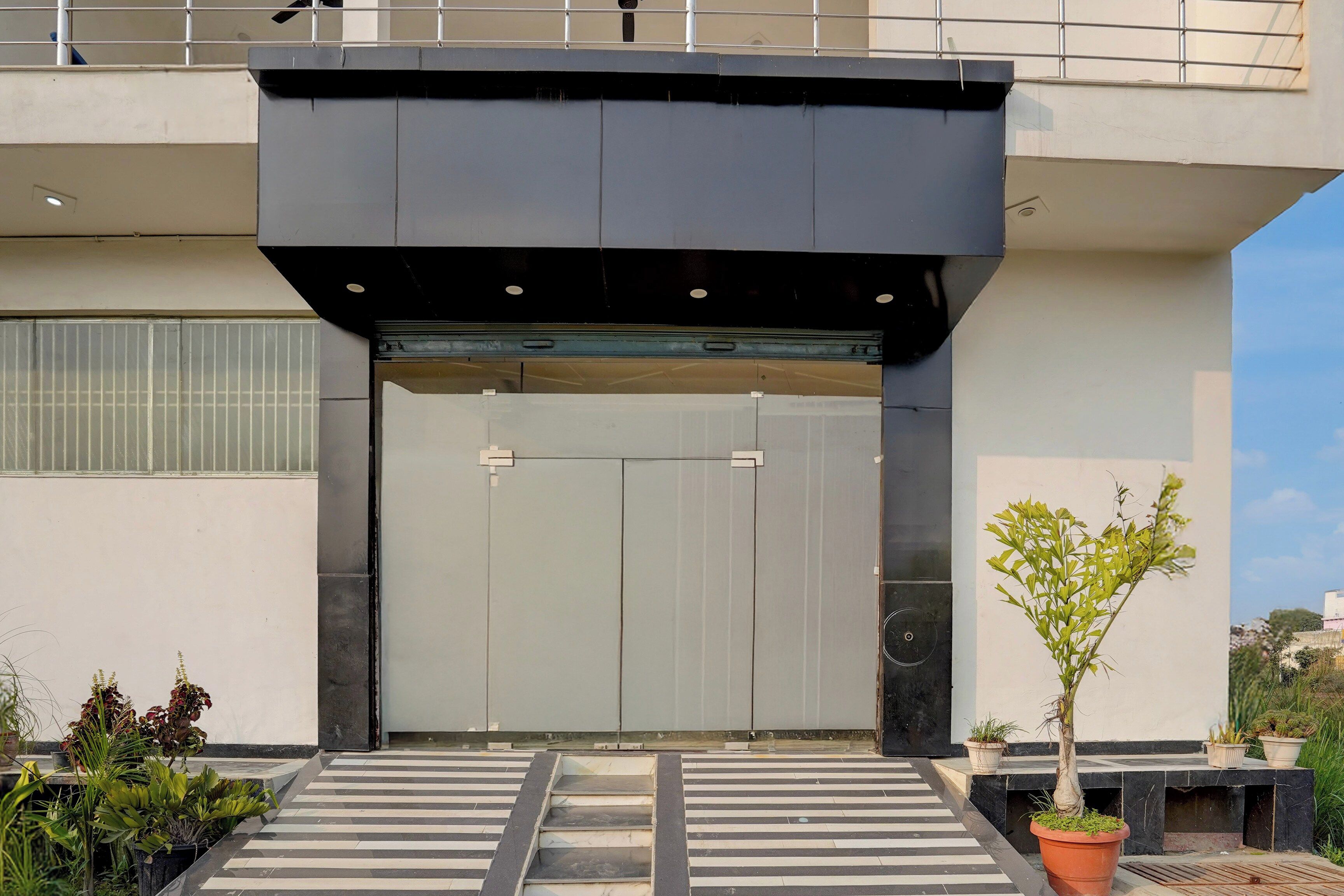 A modern building entrance with a large sliding door, a small outdoor area with potted plants, and a metal railing above.