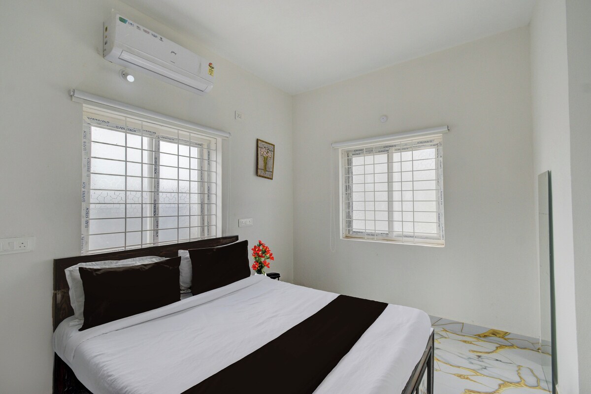 Ein Schlafzimmer mit einem Bett, zwei Fenstern mit Jalousien, einer Klimaanlage und einem am Wand montierten Lichtschalter.