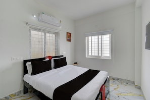 A bedroom with a bed, two windows with blinds, an air conditioning unit, and a small table with a chair. - Super Hotel O Gandimaisamma Thalli (Hyderabad)