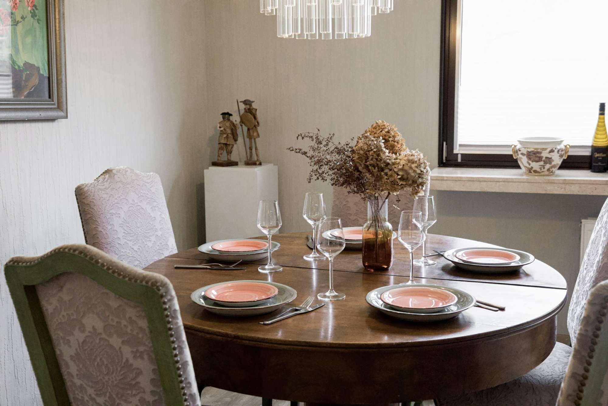 A round dining table with plates, glasses, and a vase of dried flowers.