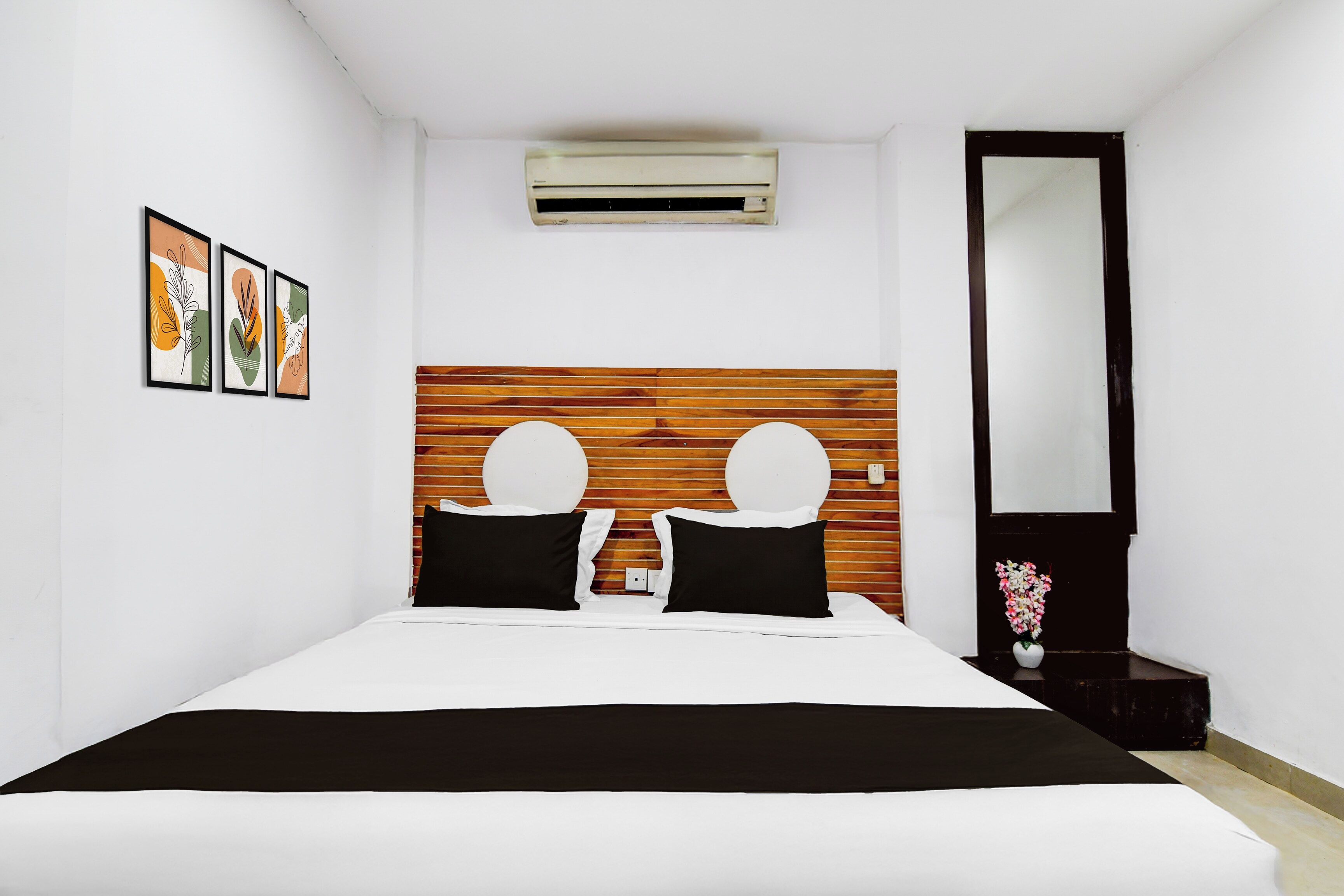 A hotel room with a bed, a wooden headboard, a wall-mounted air conditioner, a mirror, and framed artwork on the wall.