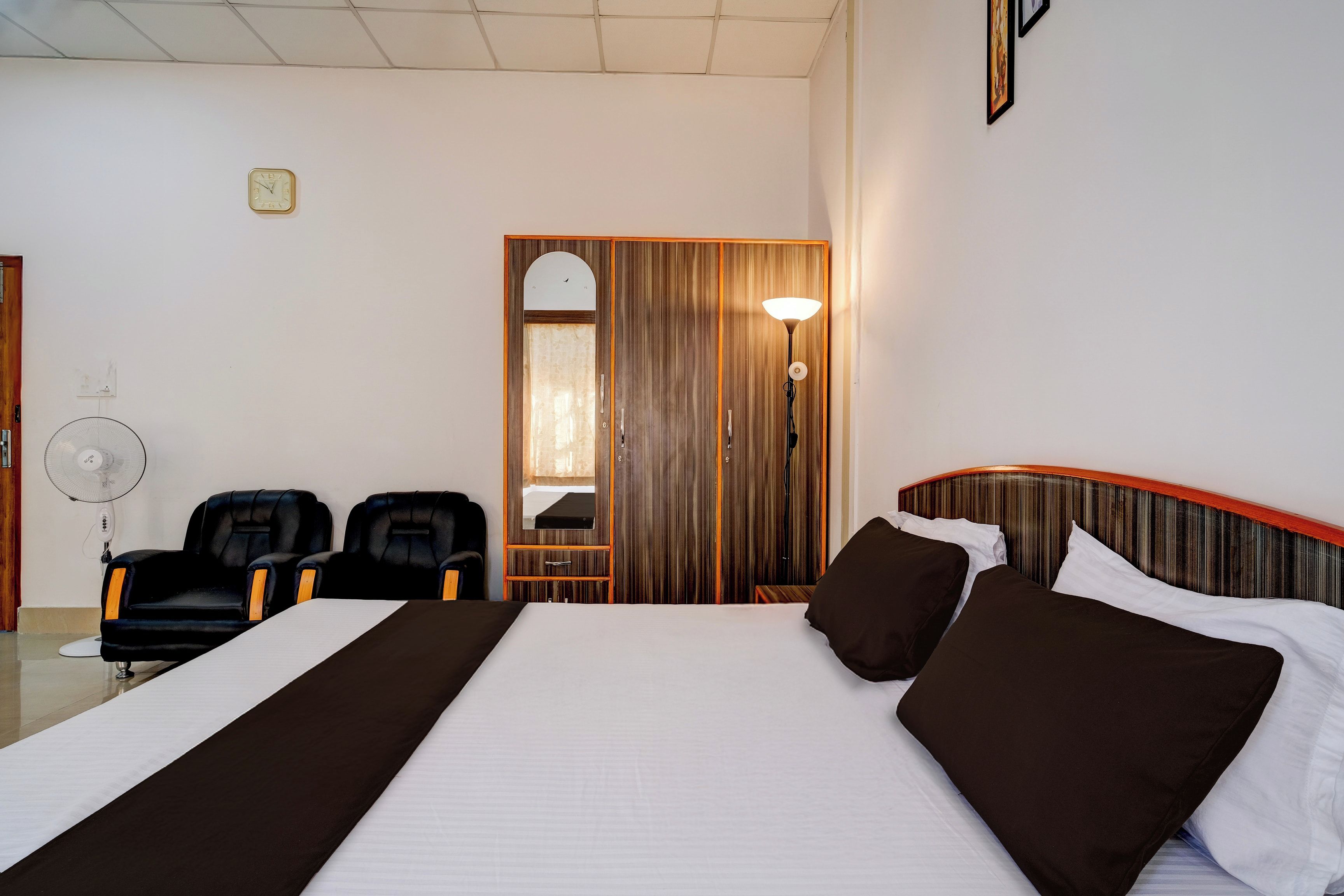 A hotel room with a large bed, two black chairs, a desk, a mirror, and a standing lamp.