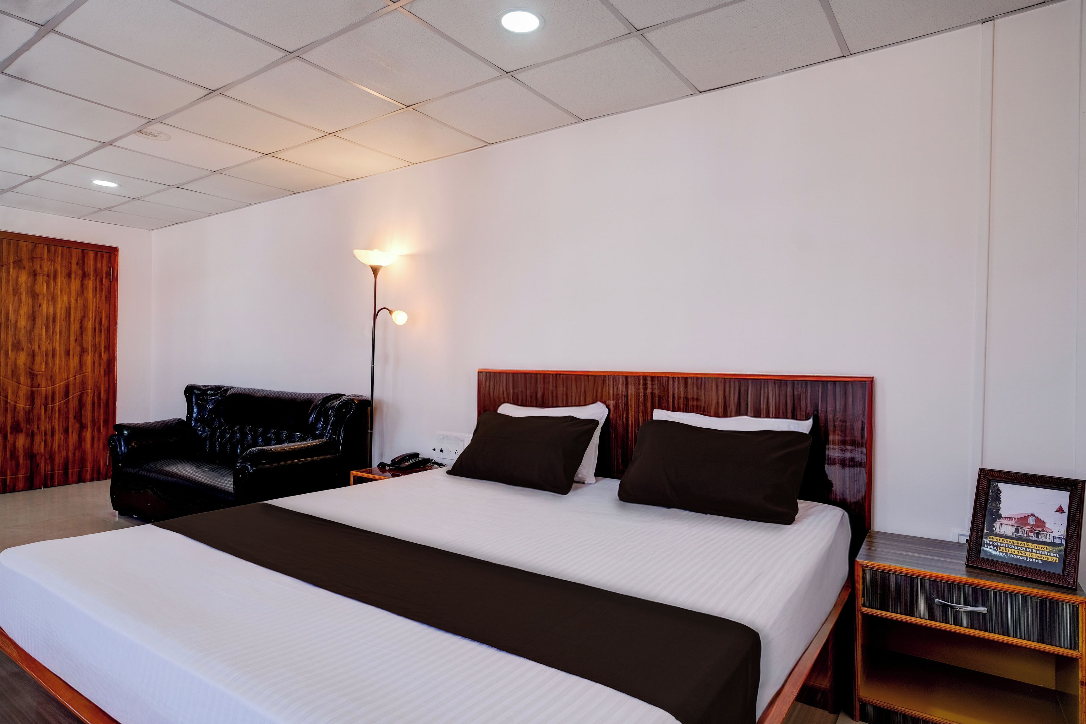 A hotel room with a large bed, a wooden headboard, a black leather sofa, a bedside table with a lamp, and a framed picture on the wall.