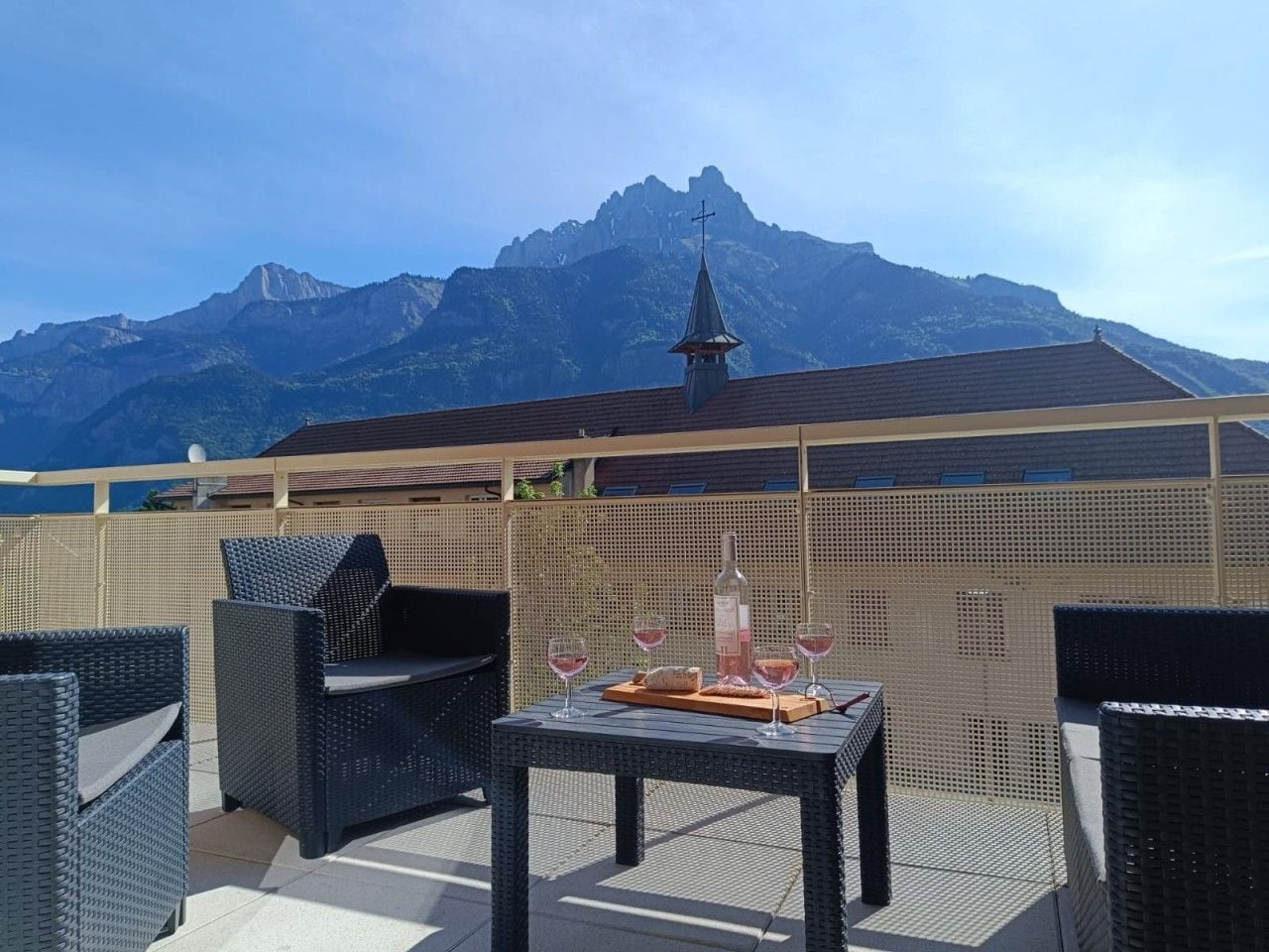 An outdoor seating area with wicker furniture, a table with a bottle and glasses, and a mountain view in the background.